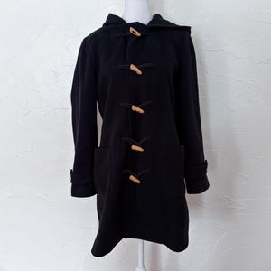 May include: A black duffel coat with a hood and wooden toggle closures. The coat has two front pockets and long sleeves with buttoned cuffs. The coat is displayed on a mannequin.