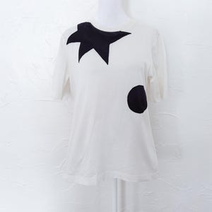 May include: White short-sleeve t-shirt with a black star and a black circle design. The star is located on the upper left chest area, and the circle is on the lower right side of the shirt. The shirt is displayed on a mannequin.