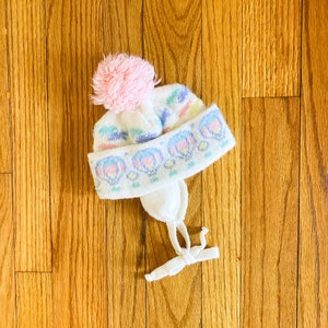 May include: White knitted hat with a pink pom pom and a colourful pattern. The hat has a tie string.
