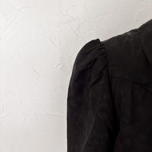 Pu&ograve; includere: Primo piano di un capo nero con un motivo strutturato. Il tessuto sembra essere un materiale leggero, forse seta o un tessuto simile. La zona delle spalle &egrave; arricciata, creando un effetto manica a sbuffo.