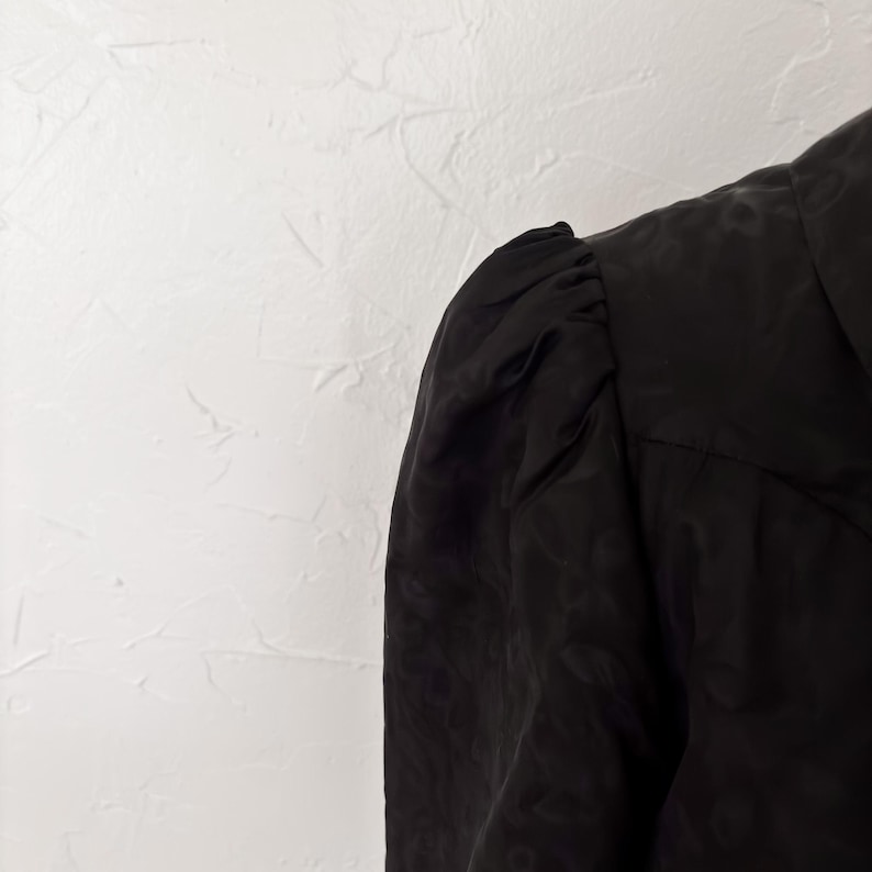 Pu&ograve; includere: Primo piano di un capo nero con un motivo strutturato. Il tessuto sembra essere un materiale leggero, forse seta o un tessuto simile. La zona delle spalle &egrave; arricciata, creando un effetto manica a sbuffo.