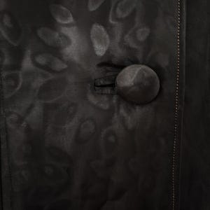 Pu&ograve; includere: Primo piano di un capo nero con un motivo strutturato e un grande bottone rotondo. Il tessuto ha una lucentezza sottile e il bottone &egrave; di un colore scuro abbinato. Una cucitura con cuciture chiare corre lungo il lato destro.
