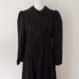 Pu&ograve; includere: Un cappotto nero vintage con colletto tondo e tre bottoni. Il cappotto ha maniche lunghe con spalle a sbuffo e un sottile motivo floreale. Il cappotto &egrave; esposto su un manichino su sfondo bianco.