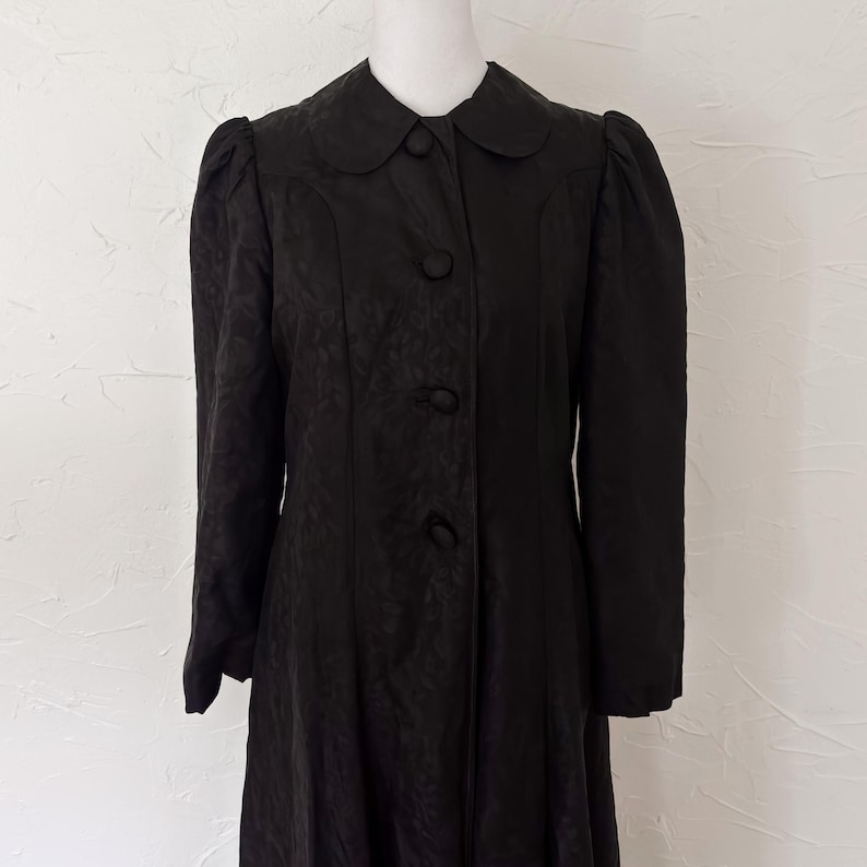 Pu&ograve; includere: Un cappotto nero vintage con colletto tondo e tre bottoni. Il cappotto ha maniche lunghe con spalle a sbuffo e un sottile motivo floreale. Il cappotto &egrave; esposto su un manichino su sfondo bianco.