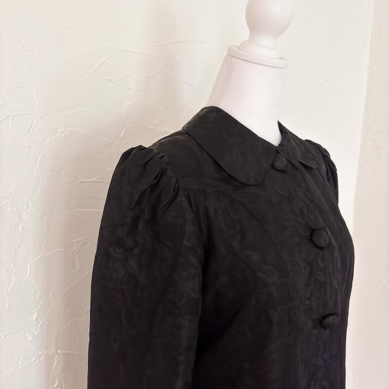 Pu&ograve; includere: Un cappotto nero vintage con colletto tondo e tre bottoni. Il cappotto ha maniche lunghe con spalle a sbuffo e un sottile motivo floreale. Il cappotto &egrave; esposto su un manichino bianco.