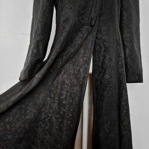 Pu&ograve; includere: Un cappotto lungo nero con un sottile motivo a foglie. Il cappotto ha una chiusura con un solo bottone e maniche lunghe. Il tessuto sembra essere un materiale liscio, forse satinato, con una leggera lucentezza. Il cappotto &egrave; aperto.