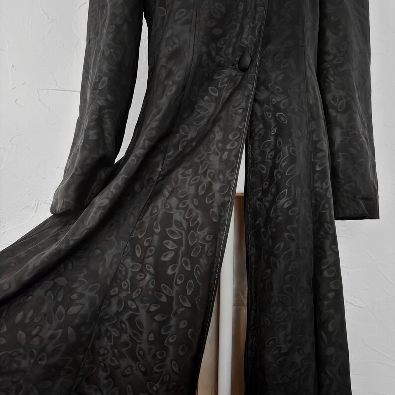Pu&ograve; includere: Un cappotto lungo nero con un sottile motivo a foglie. Il cappotto ha una chiusura con un solo bottone e maniche lunghe. Il tessuto sembra essere un materiale liscio, forse satinato, con una leggera lucentezza. Il cappotto &egrave; aperto.