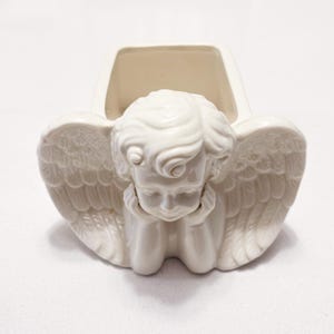 May include: Off-white ceramic angel-shaped planter. The angel has detailed wings and is depicted with its head resting on its hands. The planter has a rectangular opening at the top, suitable for small plants or decorative items.