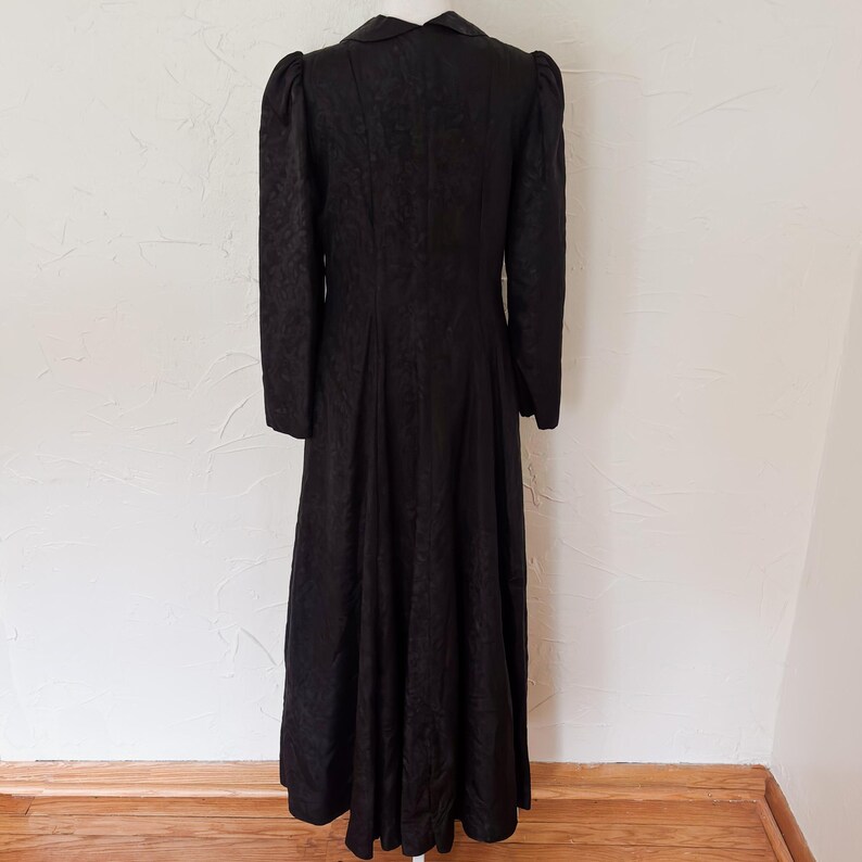 Pu&ograve; includere: Un lungo cappotto nero vintage con un motivo strutturato. Il cappotto presenta un collo, maniche lunghe e una gonna svasata. Il cappotto &egrave; esposto su un manichino su sfondo neutro.