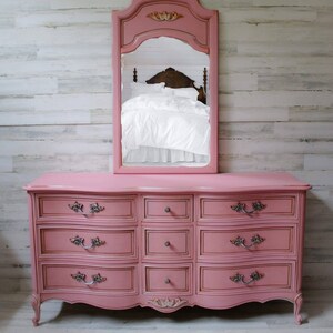 May include: A pink dresser with a curved front and eight drawers. The dresser has ornate silver handles and a carved floral design on the front. A pink mirror with a carved frame is above the dresser. The dresser is sitting on a light wood floor.