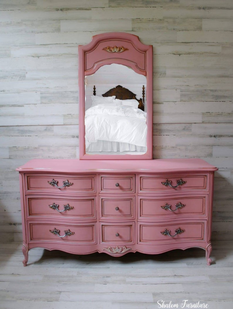 May include: A pink dresser with a curved front and eight drawers. The dresser has ornate silver handles and a carved floral design on the front. A pink mirror with a carved frame is above the dresser. The dresser is sitting on a light wood floor.