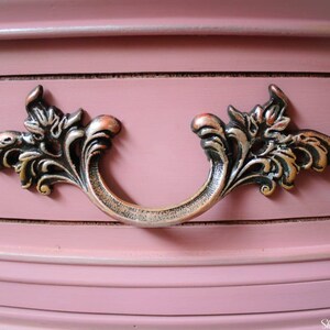 May include: A close-up of a drawer pull with a decorative, ornate design. The pull is made of metal and has a dark, aged finish. The pull is attached to a pink painted drawer.