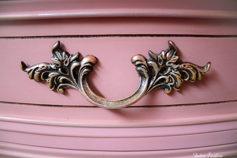 May include: A close-up of a drawer pull with a decorative, ornate design. The pull is made of metal and has a dark, aged finish. The pull is attached to a pink painted drawer.
