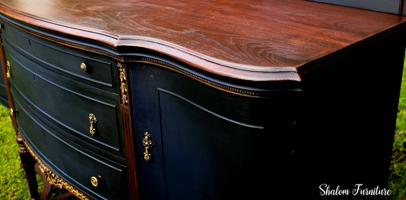 K&ouml;nnte beinhalten: Ein antiker Holz-Sideboard mit einer geschwungenen Oberseite, schwarz lackiert mit goldenen Akzenten. Das Sideboard hat drei Schubladen und eine Schrankt&uuml;r.