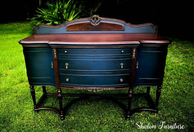 K&ouml;nnte beinhalten: Ein vintage Buffet, dunkelblau lackiert mit goldenen Akzenten und einer Mahagoni-Oberfl&auml;che. Das Buffet hat drei Schubladen und zwei T&uuml;ren. Es steht auf einem gr&uuml;nen Rasen.