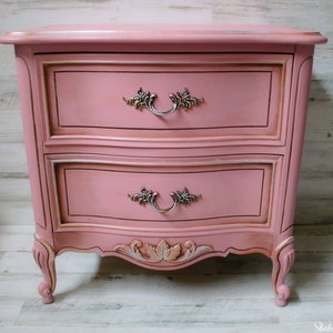 May include: A two-drawer nightstand with a pink painted finish and ornate gold accents. The drawers have silver handles with floral details. The nightstand has carved legs and a curved front.