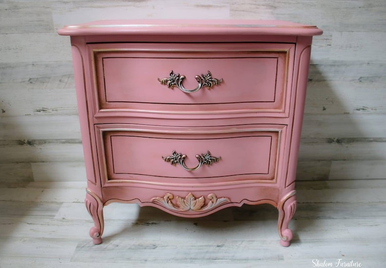 May include: A two-drawer nightstand with a pink painted finish and ornate gold accents. The drawers have silver handles with floral details. The nightstand has carved legs and a curved front.