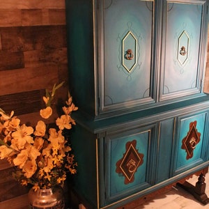 May include: A teal blue vintage hutch with gold accents and ornate brass hardware. The hutch has two upper cabinets and two lower cabinets with a wood base. The hutch is standing on a light wood floor.