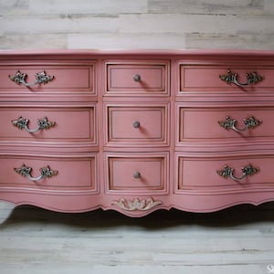 May include: A vintage-style dresser with a pink painted finish and ornate gold trim. It has nine drawers with silver-toned handles and a curved base.