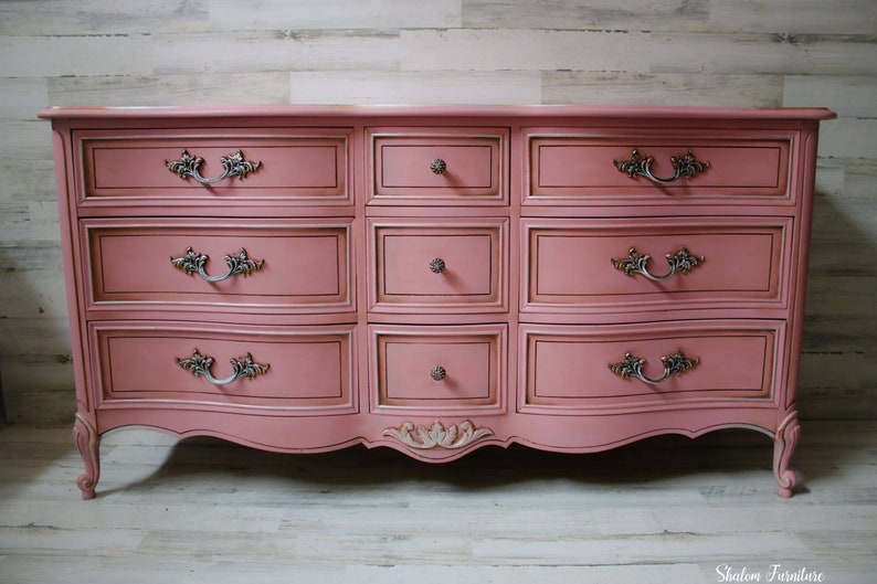 May include: A vintage-style dresser with a pink painted finish and ornate gold trim. It has nine drawers with silver-toned handles and a curved base.
