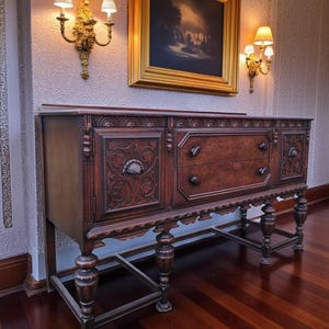 May include: A dark wood antique buffet with carved floral designs on the doors and drawers. The buffet has three drawers and two doors, and is supported by turned legs. The buffet is in a room with sconces and a framed painting.