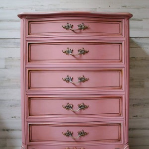 May include: A pink painted chest of drawers with ornate gold-toned hardware and a curved top. The chest has five drawers and sits on four legs. The chest is in front of a white wood panel wall. Shalom Furniture is printed in black at the bottom of the image.