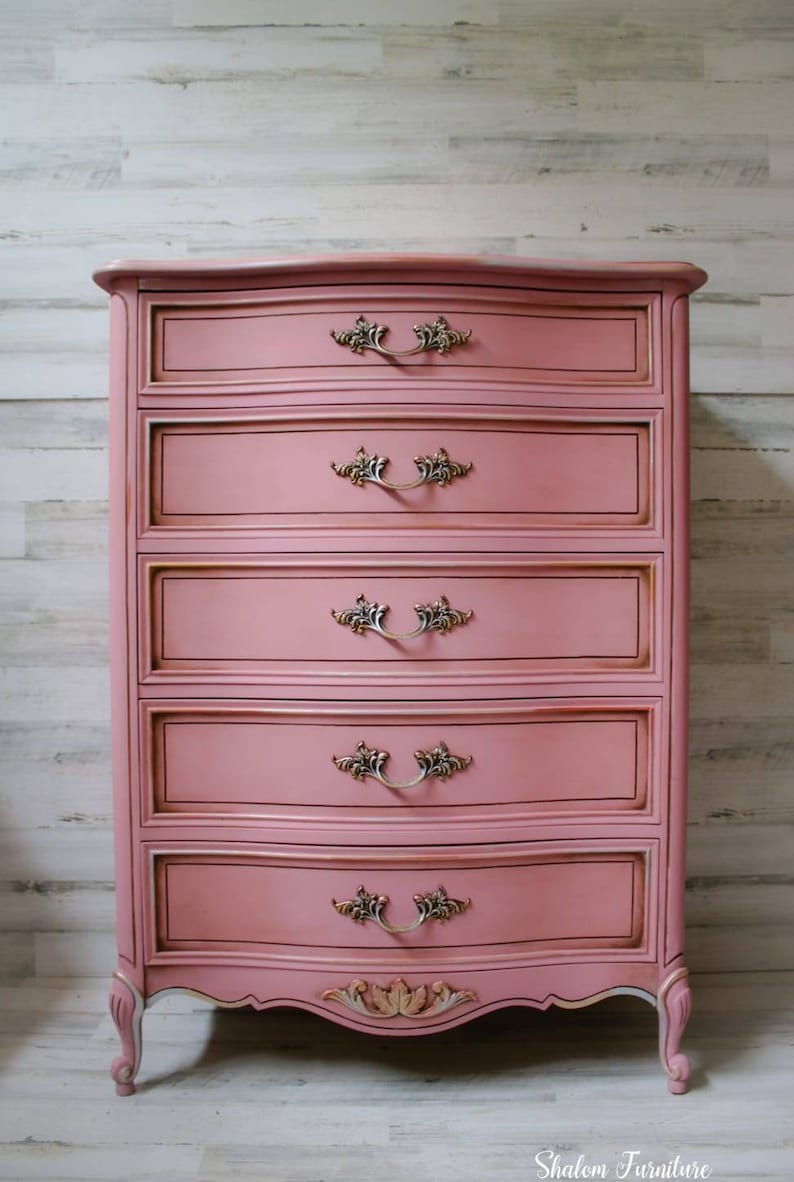 May include: A pink painted chest of drawers with ornate gold-toned hardware and a curved top. The chest has five drawers and sits on four legs. The chest is in front of a white wood panel wall. Shalom Furniture is printed in black at the bottom of the image.