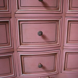 May include: A pink dresser with ornate detailing and silver drawer pulls. The dresser has a curved front and a scalloped base.