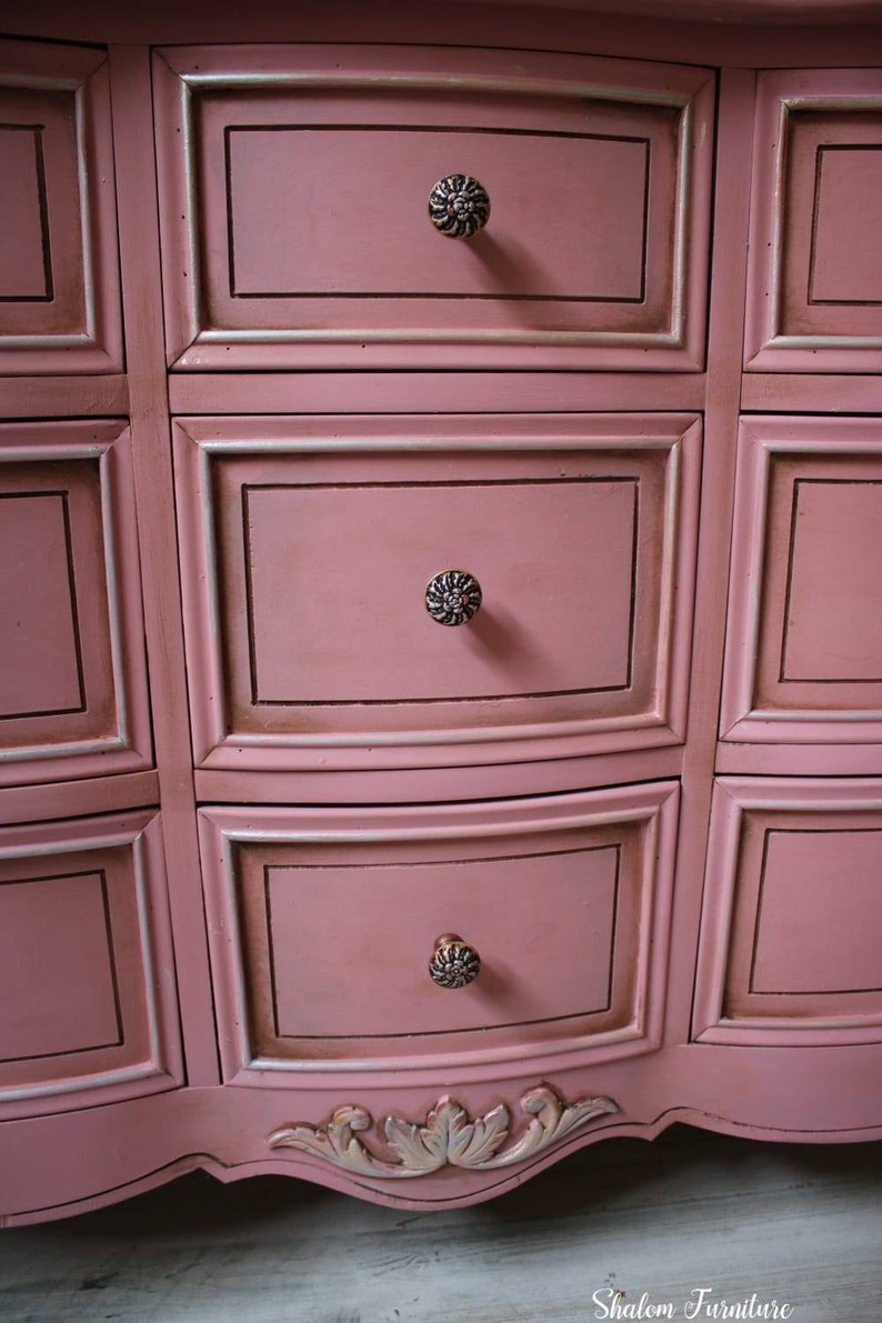 May include: A pink dresser with ornate detailing and silver drawer pulls. The dresser has a curved front and a scalloped base.