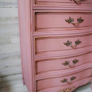 May include: A pink dresser with five drawers and ornate gold handles. The dresser has a curved bottom and sits on four legs. The dresser is painted with a distressed finish.