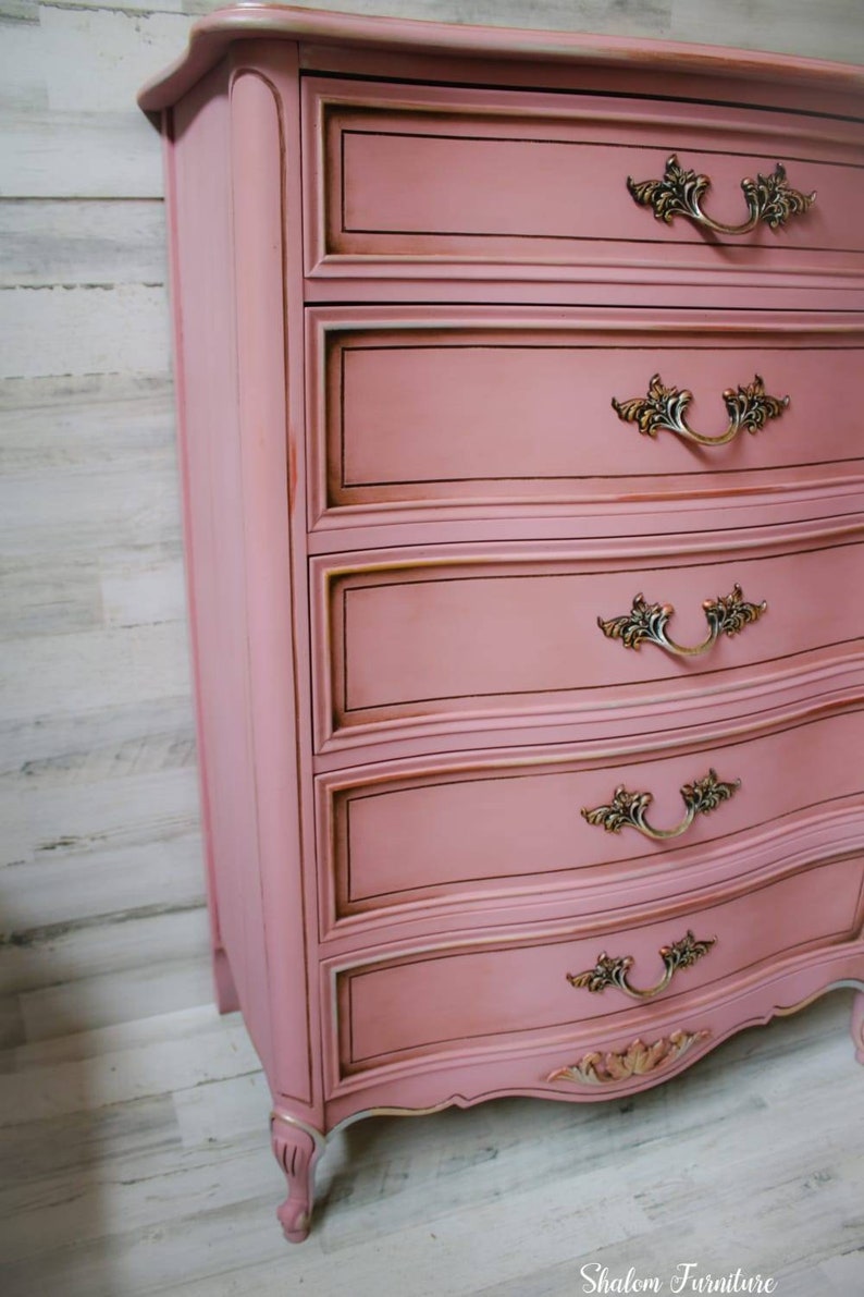 May include: A pink dresser with five drawers and ornate gold handles. The dresser has a curved bottom and sits on four legs. The dresser is painted with a distressed finish.