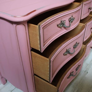 May include: A pink painted dresser with ornate details and curved drawers. The drawers are open, revealing the wooden interior. The dresser is sitting on a light wood floor.