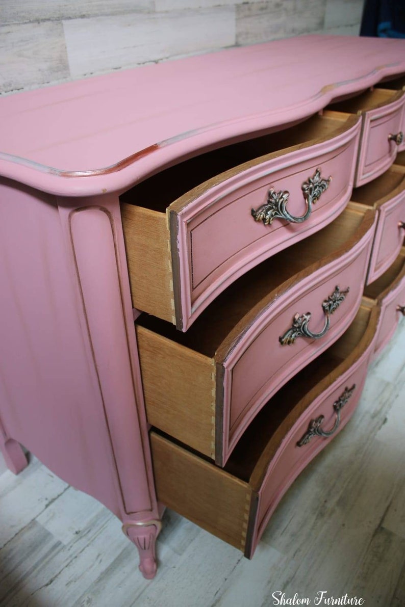 May include: A pink painted dresser with ornate details and curved drawers. The drawers are open, revealing the wooden interior. The dresser is sitting on a light wood floor.