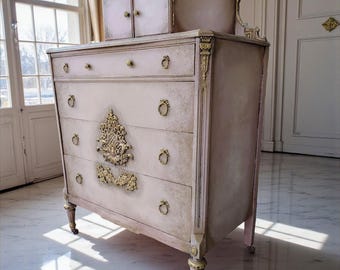 Könnte beinhalten: Eine hellrosa antike Kommode mit goldenen Akzenten. Die Kommode hat vier Schubladen mit verzierten goldenen Griffen und einem dekorativen goldenen Schmuck. Obenauf befindet sich ein kleiner Schrank. Die Kommode steht in einem Raum mit Marmorboden und Kronleuchter.