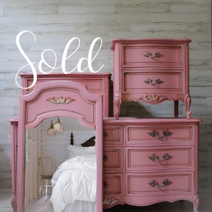 May include: A set of three pink painted vintage bedroom furniture pieces, including a dresser with four drawers, a nightstand with two drawers, and a vanity mirror with a carved frame. The furniture is decorated with ornate details and has a distressed finish. The furniture is sold.