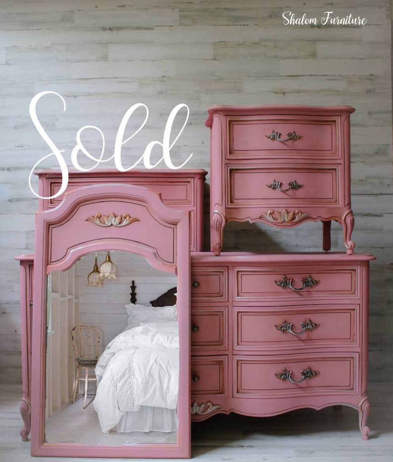 May include: A set of three pink painted vintage bedroom furniture pieces, including a dresser with four drawers, a nightstand with two drawers, and a vanity mirror with a carved frame. The furniture is decorated with ornate details and has a distressed finish. The furniture is sold.