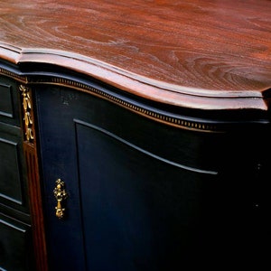 K&ouml;nnte beinhalten: Ein antiker Holz-Sideboard mit einer geschwungenen Oberseite, schwarz lackiert mit goldenen Akzenten. Das Sideboard hat drei Schubladen und eine Schrankt&uuml;r.