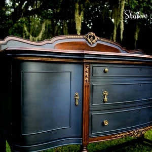 K&ouml;nnte beinhalten: Ein antiker Sideboard in Dunkelblau mit goldenen Akzenten und drei Schubladen. Das Sideboard hat eine geschwungene Front und verzierte Details. Die Oberseite des Sideboards ist aus dunklerem Holz.