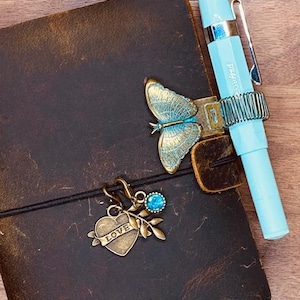 May include: A dark brown leather journal featuring a brass butterfly pen holder with a light blue fountain pen. A black elastic band secures the journal, adorned with a brass heart charm engraved with the word LOVE, a small leaf, and a blue gem.
