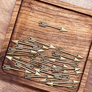 May include: A collection of small, gold-colored arrow charms arranged on a wooden tray. The arrows have detailed feather fletching and pointed tips. The tray is a warm brown color, and the arrows are scattered across its surface.
