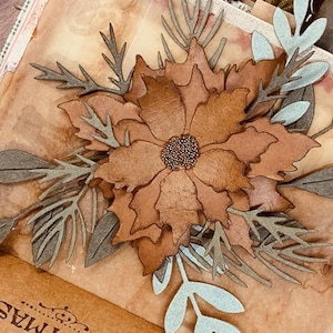 May include: A handmade paper flower with brown petals and a textured center, surrounded by gray and light blue leaves and sprigs. The flower is the focal point of a scrapbook page.