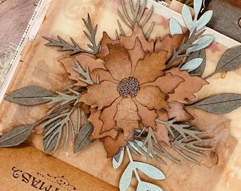 May include: A handcrafted paper flower with brown petals and a textured centre, surrounded by grey and light blue leaves and sprigs. The flower is the focal point of a scrapbook page.