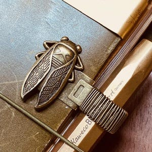 May include: A bronze-coloured cicada-shaped metal clasp and a pen with the text "Kaweco Germany". The pen is secured with a wire-wrapped band. The items rest on a leather-bound notebook with a brass-coloured bookmark.