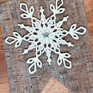 Peut inclure: Décoration de flocon de neige blanc avec une étoile au centre, fixée sur une bannière en liège. Le flocon de neige présente des détails découpés complexes et un léger effet scintillant. La bannière a un aspect rustique et texturé, avec un motif de liège naturel et un lavis bleu-vert.