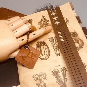 May include: A brown leather journal with a decorative metal clasp and a brown leather bookmark with a metal charm. The journal has a vintage look and feel, with a distressed paper cover and a variety of decorative elements. The journal is open to a page with a variety of decorative elements, including a brown leather bookmark with a metal charm.