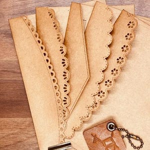 May include: A collection of tan paper envelopes with decorative scalloped edges and a metal clip. A small, square, brown leather tag with a chain is also included. The envelopes are arranged on a wooden surface.