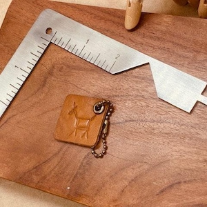 May include: A stainless steel square ruler with inch markings and a unique shape, alongside a small, square, brown leather keychain with a deer design. The items rest on a wooden surface.
