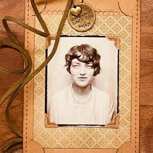May include: A vintage-style photo tag featuring a sepia-toned portrait of a woman. The tag is made of tan-colored, stitched paper with a patterned background. A small, gold-colored charm with text and a heart hangs from a green cord.