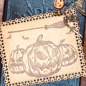 May include: A Halloween-themed journal with a decorative charm. The journal is dark blue with embossed text. A decorative card with three carved pumpkin illustrations and a broom is attached. Charms include a pumpkin, owl, and sword.
