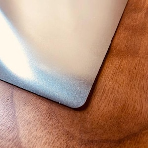 May include: A corner of a silver metal sheet with a rounded edge, set against a wooden surface. The metal has a slightly textured appearance, reflecting light. The wood grain is visible in the background.